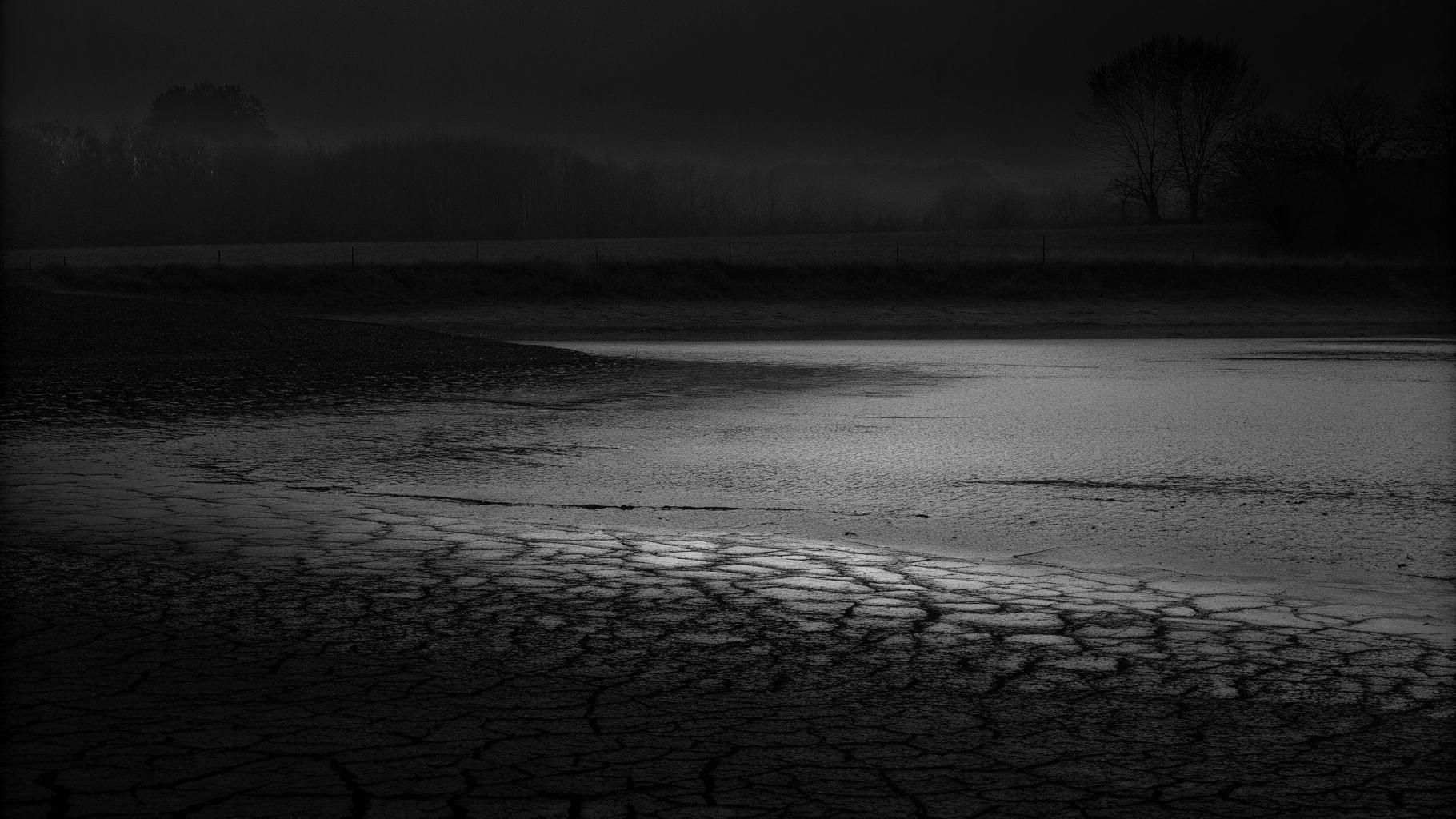 Paysage rural de la Manche avec sol craquelé et rivière à faible débit sous un ciel pâle.
