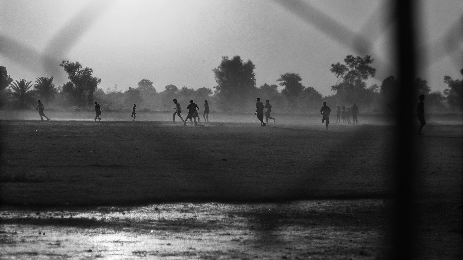 Illustration abstraite d'un match de football intense sous une lumière ardente, avec des joueurs en mouvement floutés, sans texte, sans logo, sans visage reconnaissable, couleurs sobres, format paysage 3:2